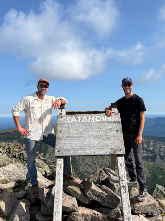 Jake and Ben standing with a sign at the top of a mountain